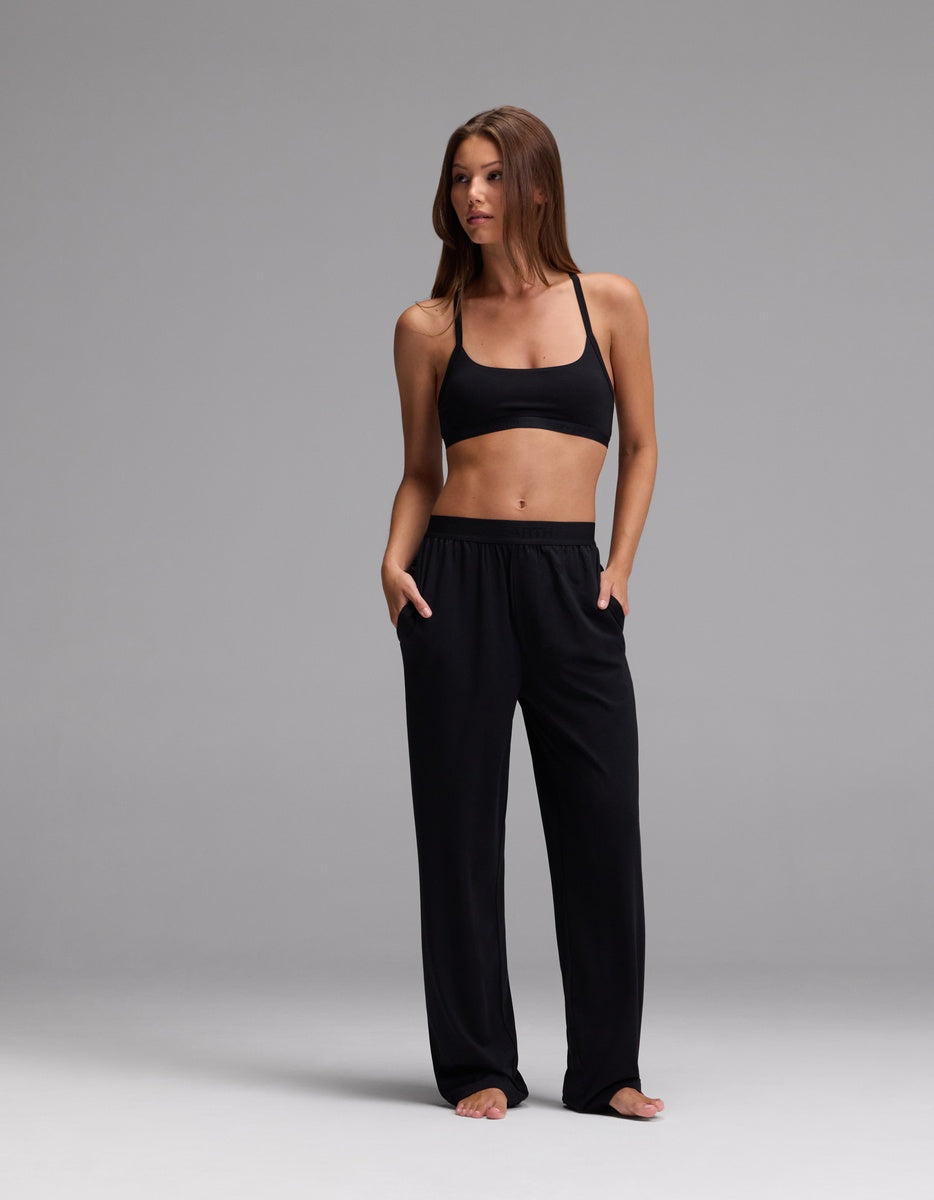 A woman stands barefoot against a gray background, wearing a black sports bra and Cozy Earth Women’s First Layer Pajama Pant with her hands in the pockets, looking to the side.