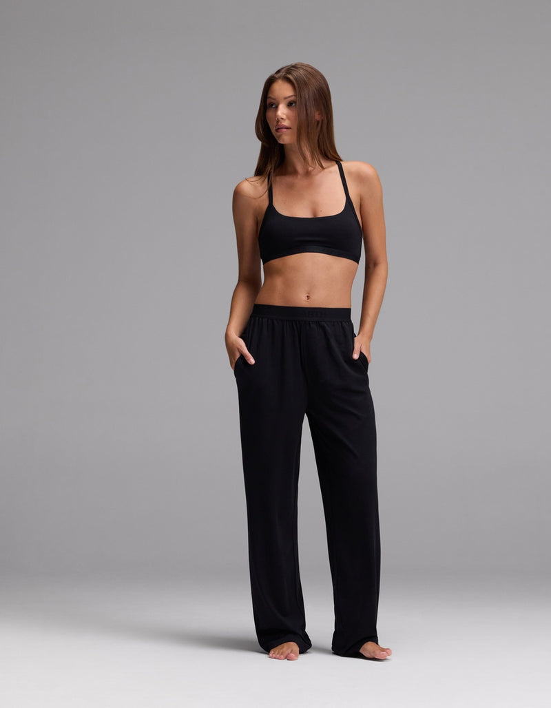 A woman stands barefoot against a gray background, wearing a black sports bra and Cozy Earth Women’s First Layer Pajama Pant with her hands in the pockets, looking to the side.