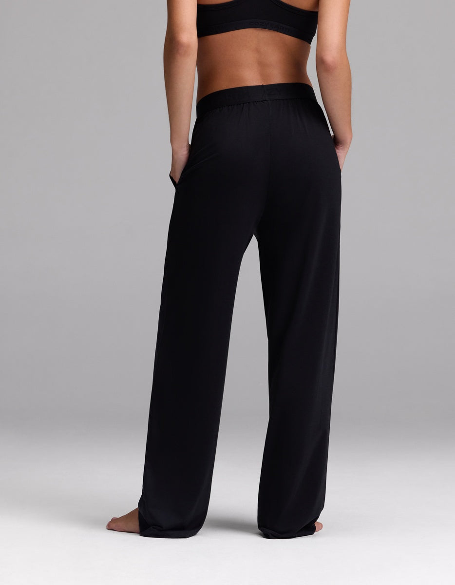 A woman models the Cozy Earth Women’s First Layer Pajama Pant in black, barefoot and facing away from the camera against a plain gray background.