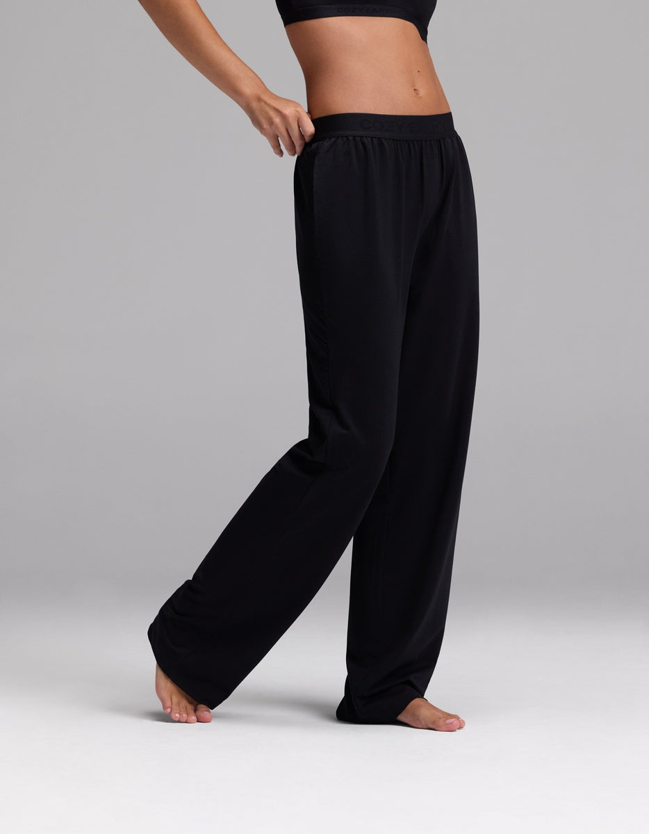 A person wearing Cozy Earth Women’s First Layer Pajama Pant in Jet Black stands barefoot on a light gray surface, posing with one hand on their hip and one leg slightly bent.