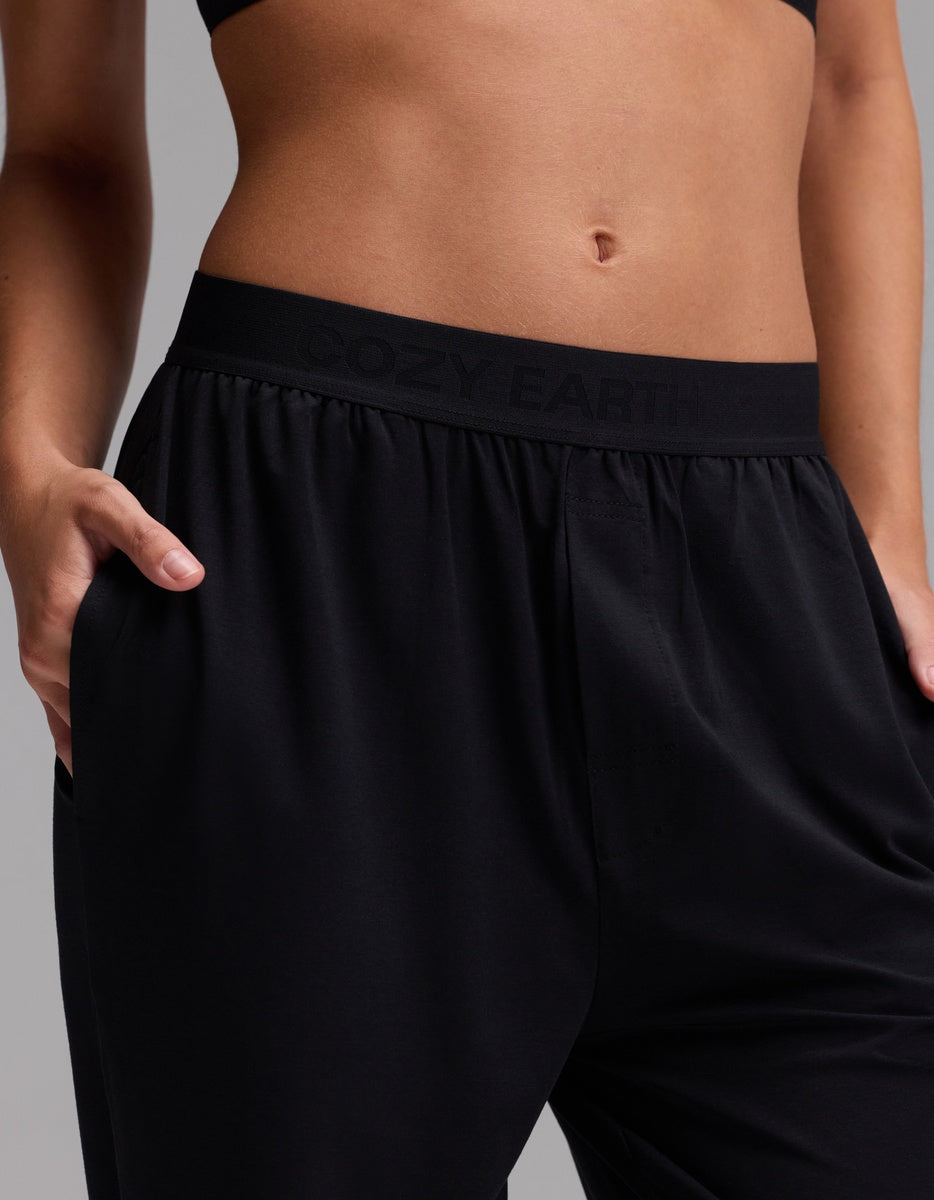 A person wearing Cozy Earth’s Women’s First Layer Pajama Pant in black, with hands in the pockets, paired with a black sports bra. The pants feature a waistband that displays “COZY EARTH,” and the background is plain and neutral.
