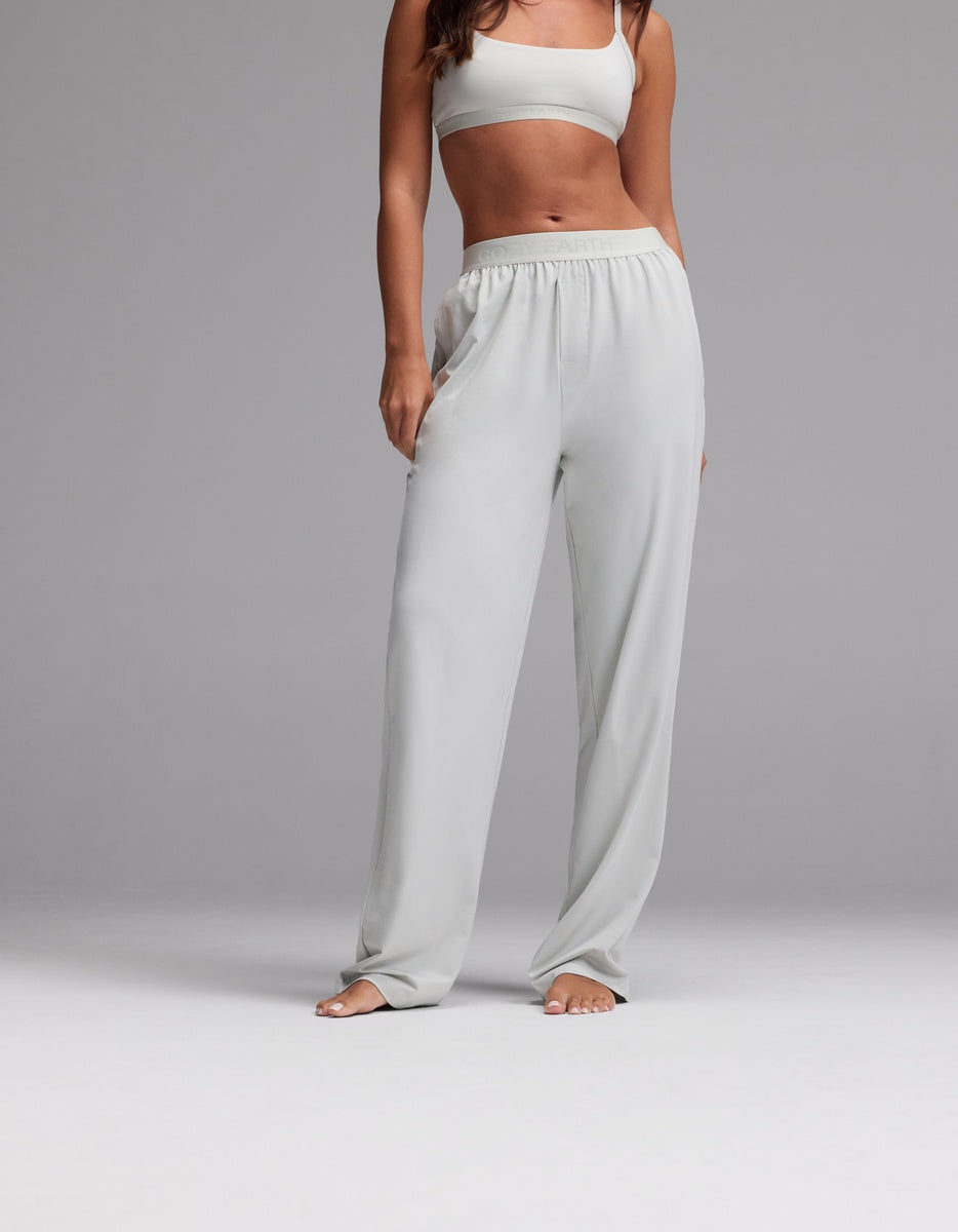 A woman wearing Cozy Earth’s Women’s First Layer Pajama Pant in Laurel stands barefoot against a plain gray background, with her hands in her pockets and her head cropped out of the image.|Color:Laurel
