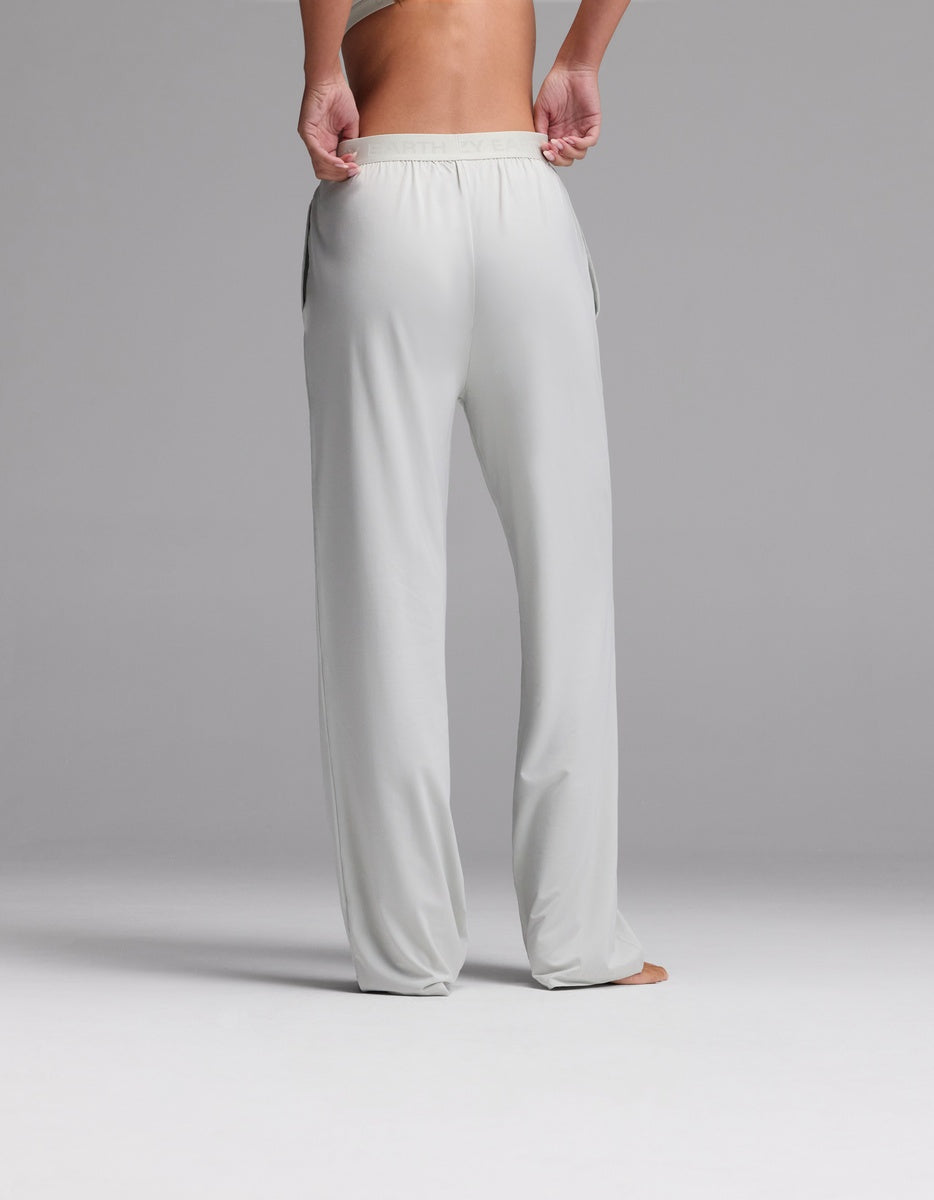 A person stands barefoot on a gray surface, shown from the waist down and back view, wearing Cozy Earth’s Women’s First Layer Pajama Pant in light gray—a loose-fitting style with an elastic waistband and their hands in the pockets.