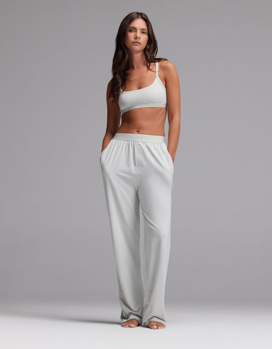 A woman stands barefoot against a plain gray background, wearing a light gray sports bra and Cozy Earth Women’s First Layer Pajama Pants—loose, high-waisted lounge pants with her hands in the pockets.