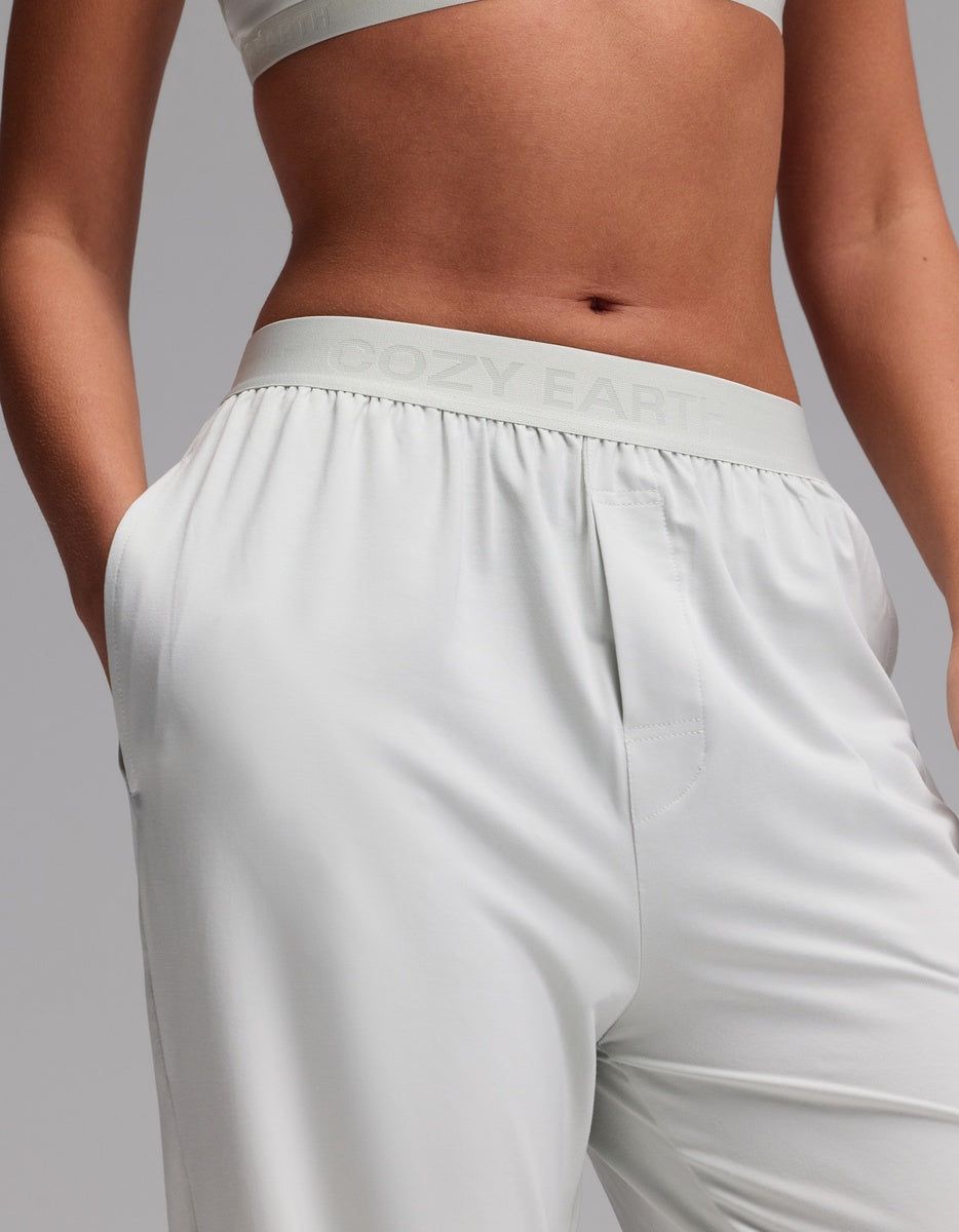 A person stands with hands in the pockets of light gray Women’s First Layer Pajama Pants by Cozy Earth, featuring a branded waistband and paired with a matching crop top.
