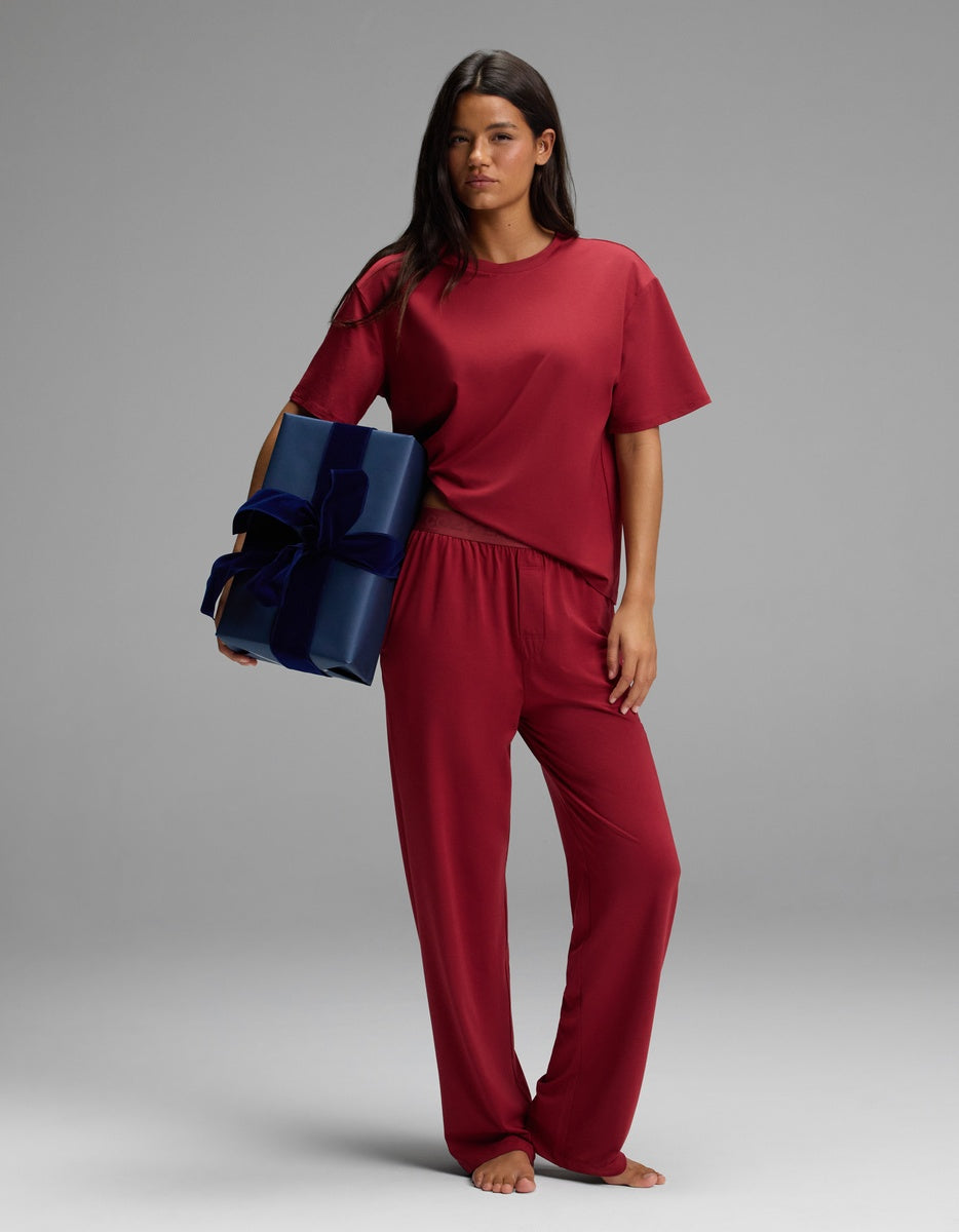 A woman wearing a matching dark red short-sleeved top and Women’s First Layer Pajama Pant, holding a dark blue gift box with a velvet ribbon.