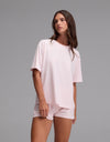A woman with long brown hair wears the Cozy Earth Women’s First Layer Pajama Short Sleeve Tee and matching shorts in light pink, standing against a plain gray background while gazing slightly to the side.|Color:Ballet
