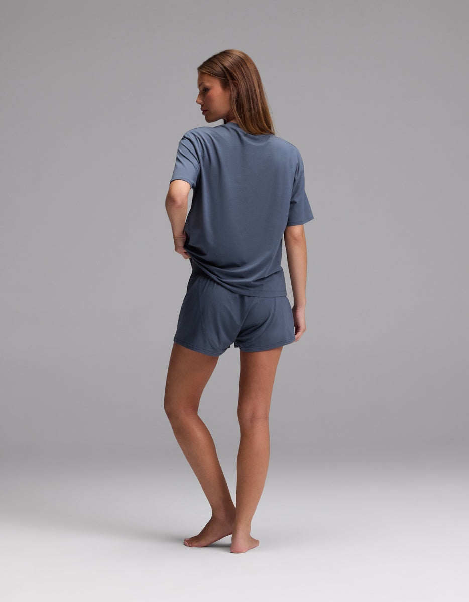 A woman, barefoot and facing away, models the Cozy Earth Women’s First Layer Pajama Short Sleeve Tee & Boxer Short Set in gray against a plain light gray background.|Color:Dawn
