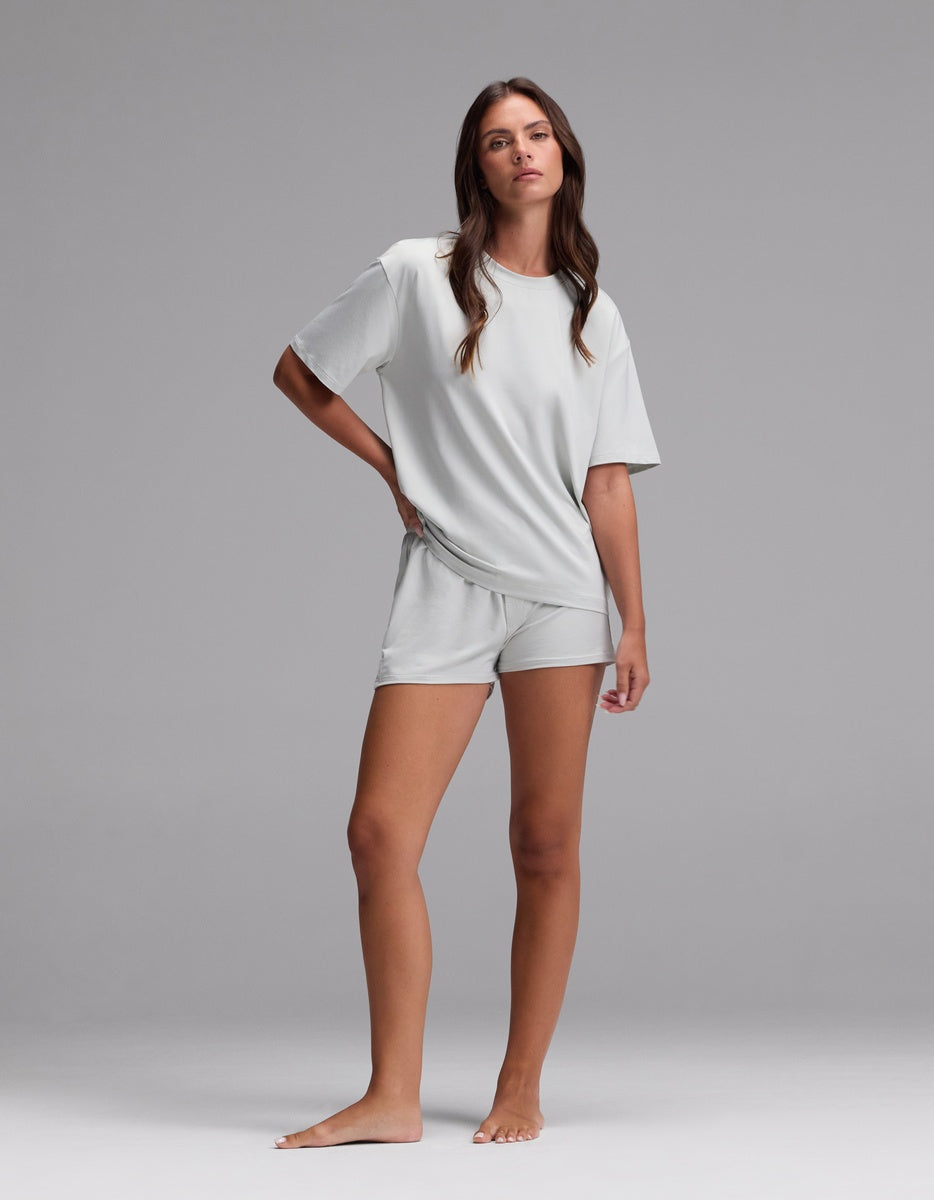 A woman stands barefoot against a plain background, wearing the Women’s First Layer Pajama Short Sleeve Tee & Boxer Short Set by Cozy Earth in Laurel, with a relaxed pose and neutral expression.|Color:Laurel