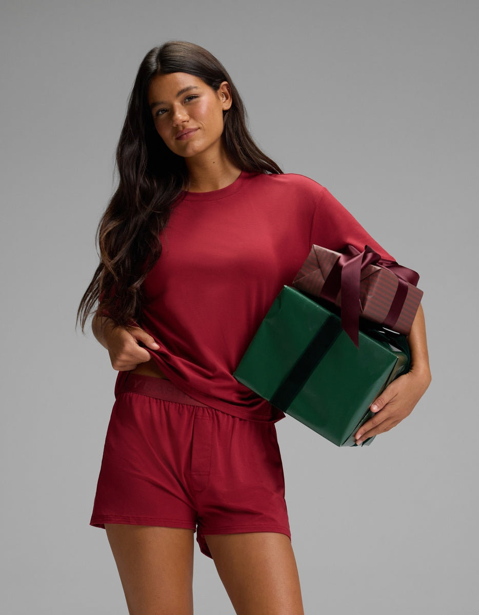 A person wearing a burgundy Women’s First Layer Pajama Short Sleeve Tee & Boxer Short Set by Cozy Earth, holding two wrapped gifts.