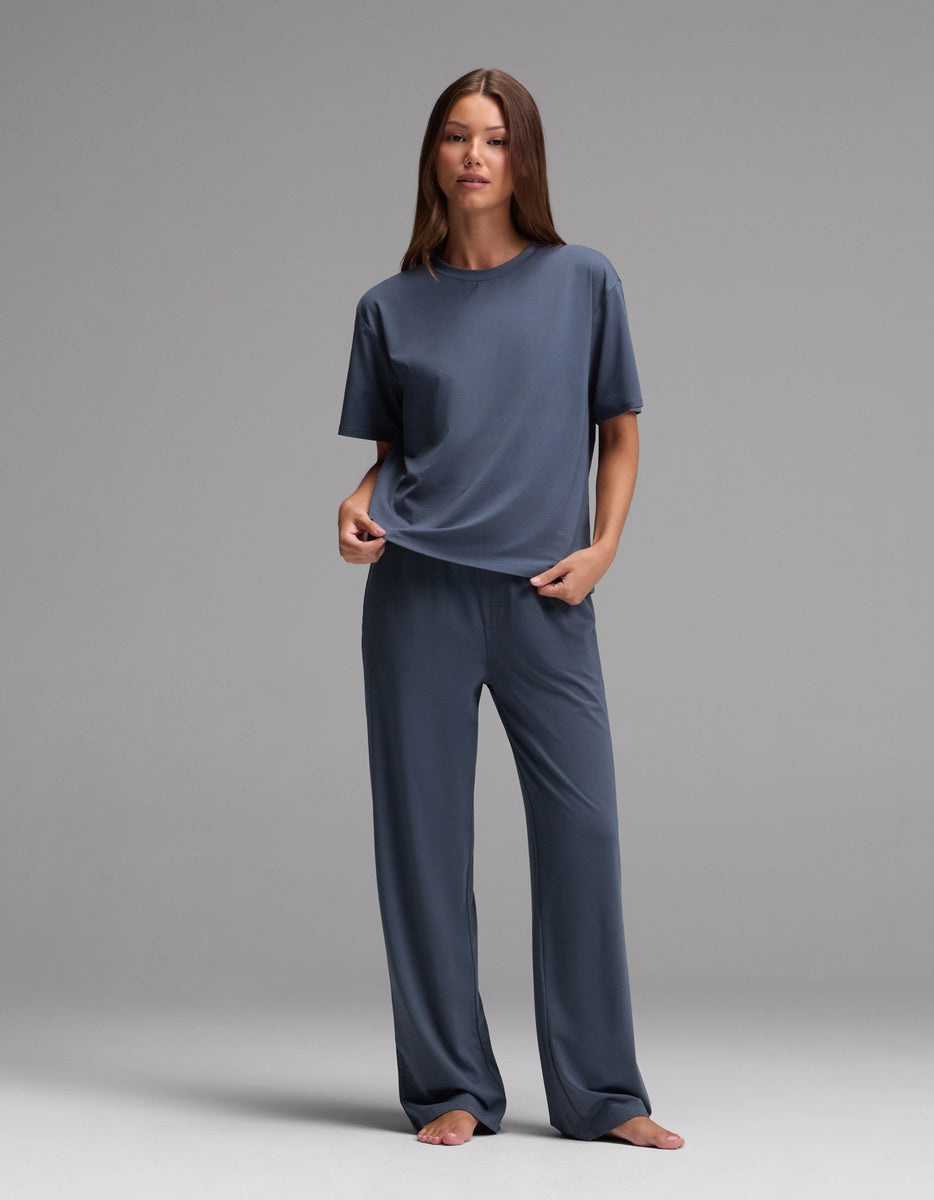 A woman stands barefoot against a gray background, wearing the Cozy Earth Women’s First Layer Pajama Short Sleeve Tee in blue along with matching wide-leg pants. She holds part of her shirt with one hand and looks straight ahead.