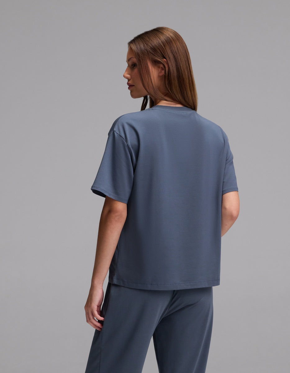A person with straight brown hair is standing and facing away from the camera, wearing the Cozy Earth Women’s First Layer Pajama Short Sleeve Tee in a loose blue-gray style, paired with matching pants against a plain gray background.