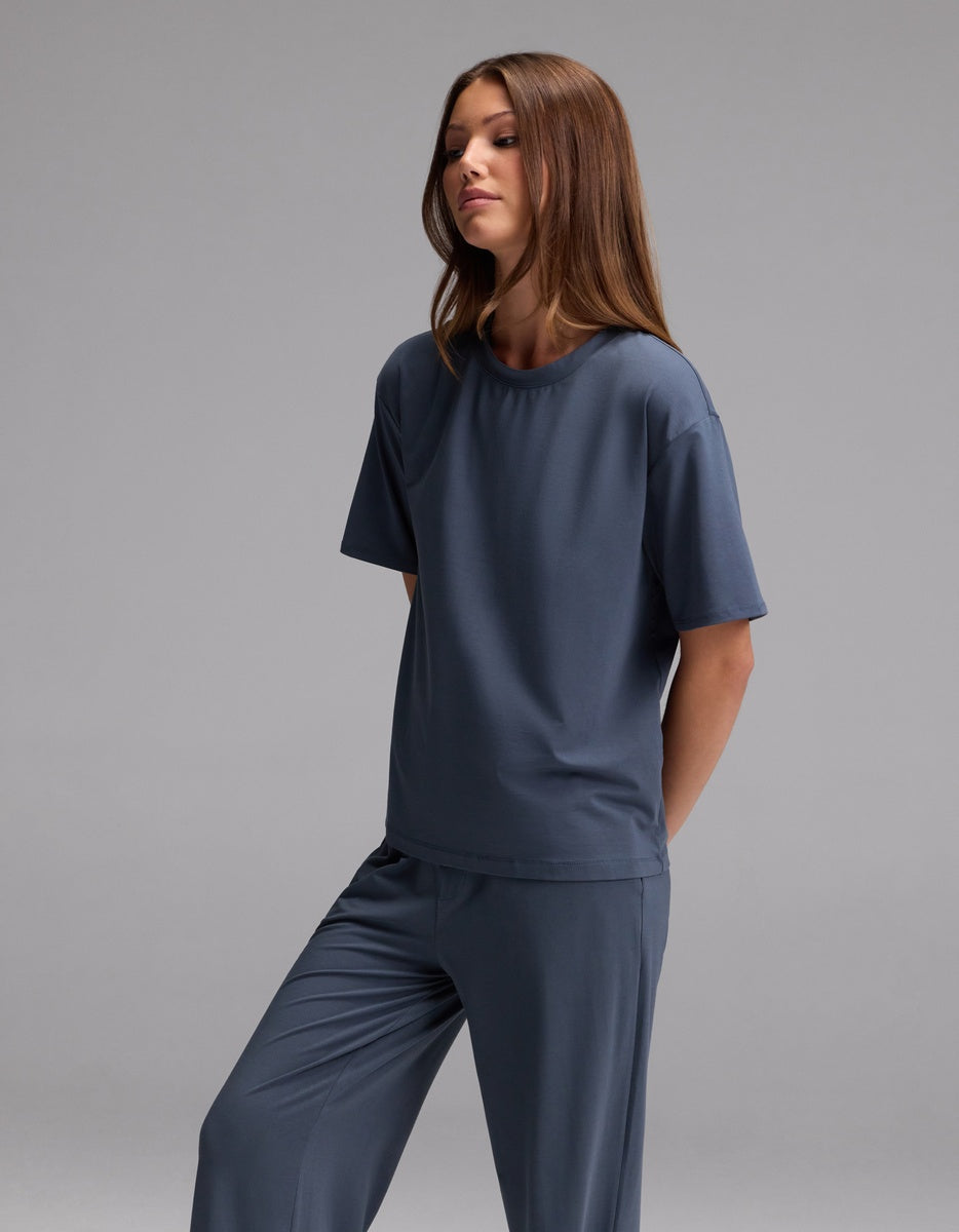 A woman with long brown hair stands against a plain gray background, wearing the Cozy Earth Women’s First Layer Pajama Short Sleeve Tee in dark blue along with matching pants. She gazes slightly to the side with a relaxed expression.