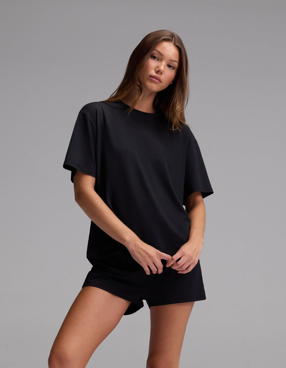 A woman wearing the Cozy Earth Women’s First Layer Pajama Short Sleeve Tee and matching black shorts stands against a plain gray background, looking at the camera with a relaxed pose and neutral expression.
