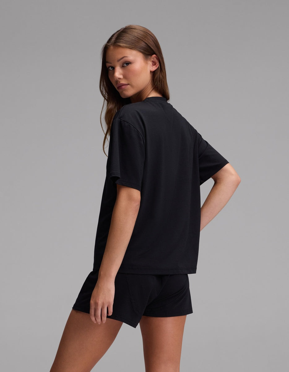 A woman with long brown hair stands sideways, looking over her shoulder. She is wearing the Cozy Earth Women’s First Layer Pajama Short Sleeve Tee in black, paired with matching black shorts, against a plain gray background.