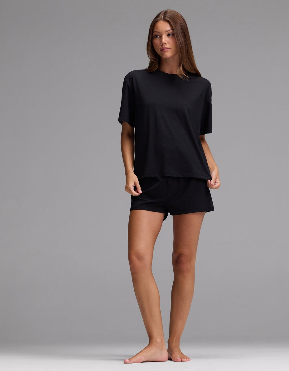 A woman stands barefoot against a gray background, wearing Cozy Earth’s Women’s First Layer Pajama Short Sleeve Tee in black, paired with matching black shorts. She has long, straight brown hair and a neutral expression, holding the hem of her shirt with her right hand.