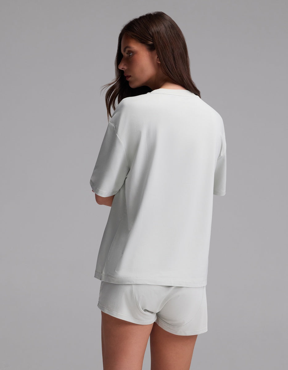 A woman with long brown hair stands facing away, wearing the Cozy Earth Women’s First Layer Pajama Short Sleeve Tee in white along with matching white shorts, set against a plain gray background.