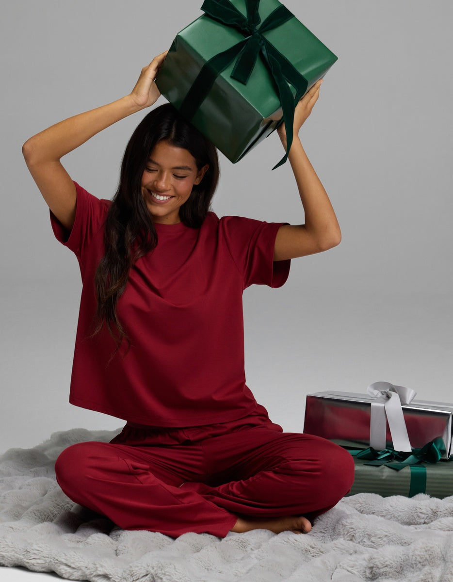 A smiling woman wearing a dark red Women’s First Layer Pajama Short Sleeve Tee & Pajama Pant Set sits cross-legged on a gray faux fur rug while holding a green gift box above her head.