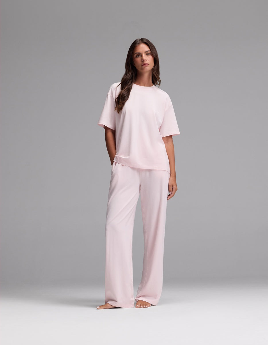 A woman stands barefoot against a plain gray background, wearing the Cozy Earth Women’s First Layer Pajama Short Sleeve Tee & Pajama Pant Set in Ballet—a loose-fitting, light pink ensemble featuring short sleeves and wide-leg pants.