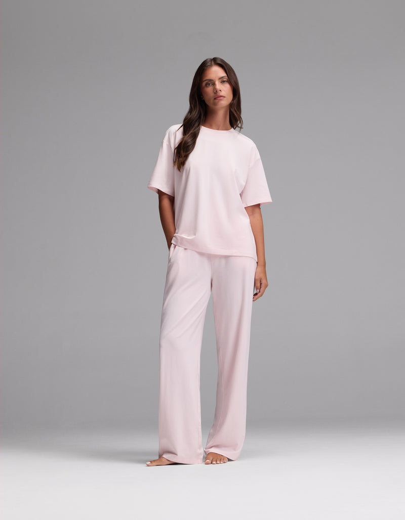 A woman stands barefoot against a plain gray background, wearing the Cozy Earth Women’s First Layer Pajama Short Sleeve Tee & Pajama Pant Set in Ballet—a loose-fitting, light pink ensemble featuring short sleeves and wide-leg pants.