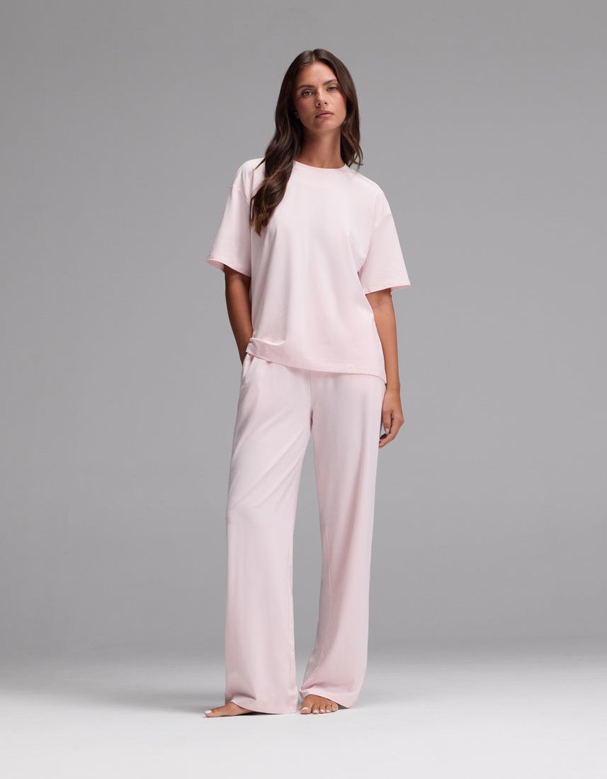 A woman stands barefoot against a plain gray background, wearing the Cozy Earth Women’s First Layer Pajama Short Sleeve Tee & Pajama Pant Set in Ballet—a loose-fitting, light pink ensemble featuring short sleeves and wide-leg pants.|Color:Ballet