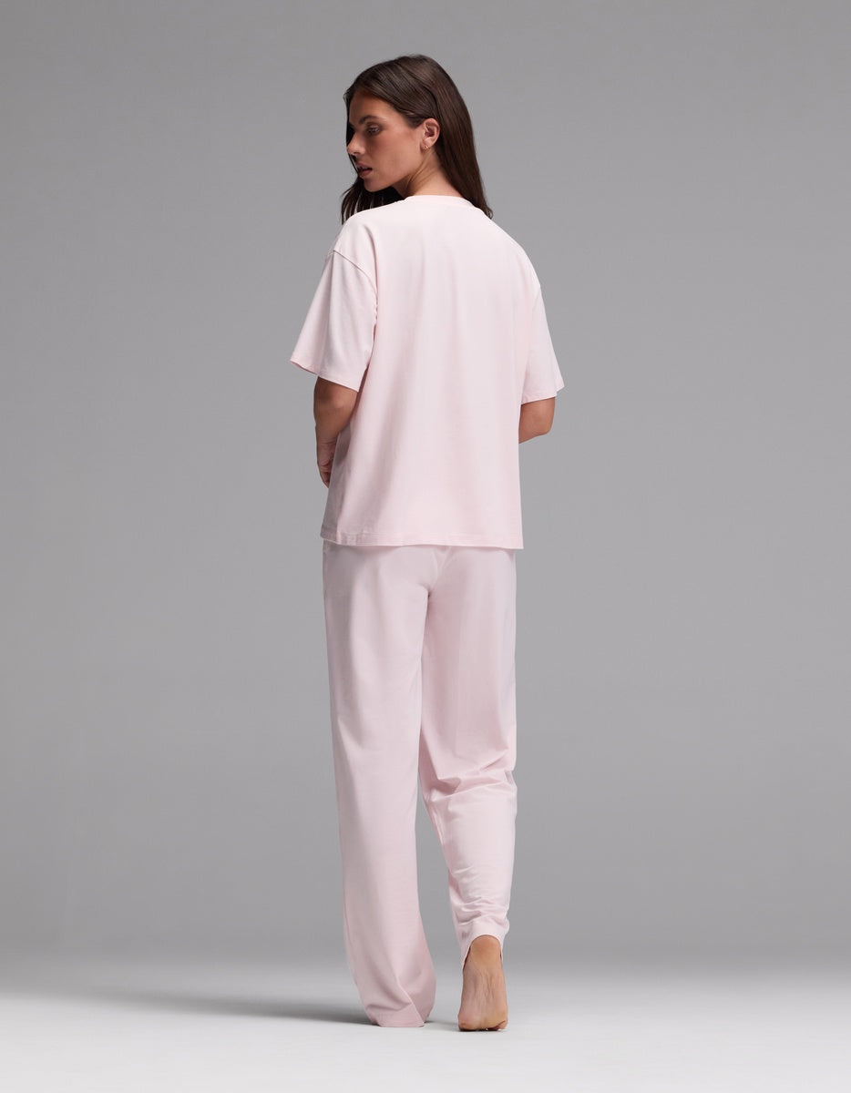 A person stands barefoot on a gray background, facing away from the camera and looking over their shoulder, wearing the Cozy Earth Women’s First Layer Pajama Short Sleeve Tee & Pajama Pant Set in light pink.
