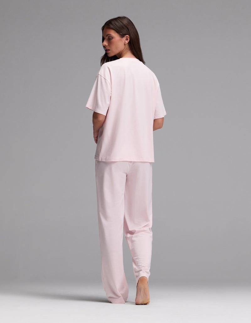 A person stands barefoot on a gray background, facing away from the camera and looking over their shoulder, wearing the Cozy Earth Women’s First Layer Pajama Short Sleeve Tee & Pajama Pant Set in light pink.