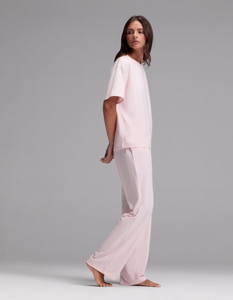 A woman with long brown hair wears the Cozy Earth Women’s First Layer Pajama Short Sleeve Tee & Pajama Pant Set in light pink. She stands barefoot on a plain gray background, looking over her shoulder with a neutral expression.