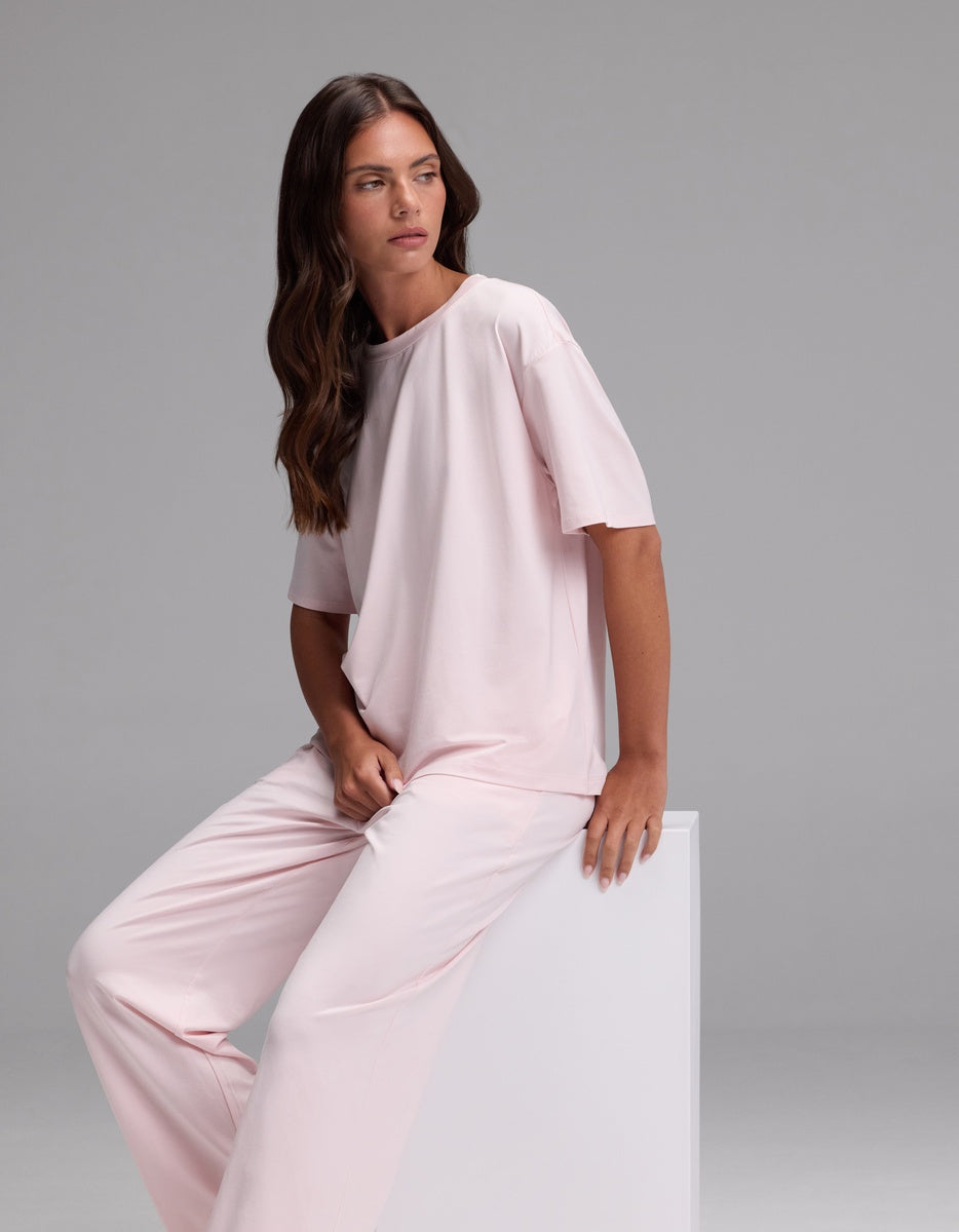 A woman with long brown hair is wearing the Cozy Earth Women’s First Layer Pajama Short Sleeve Tee & Pajama Pant Set in light pink as she sits on a white block, looking to the side against a plain gray background.