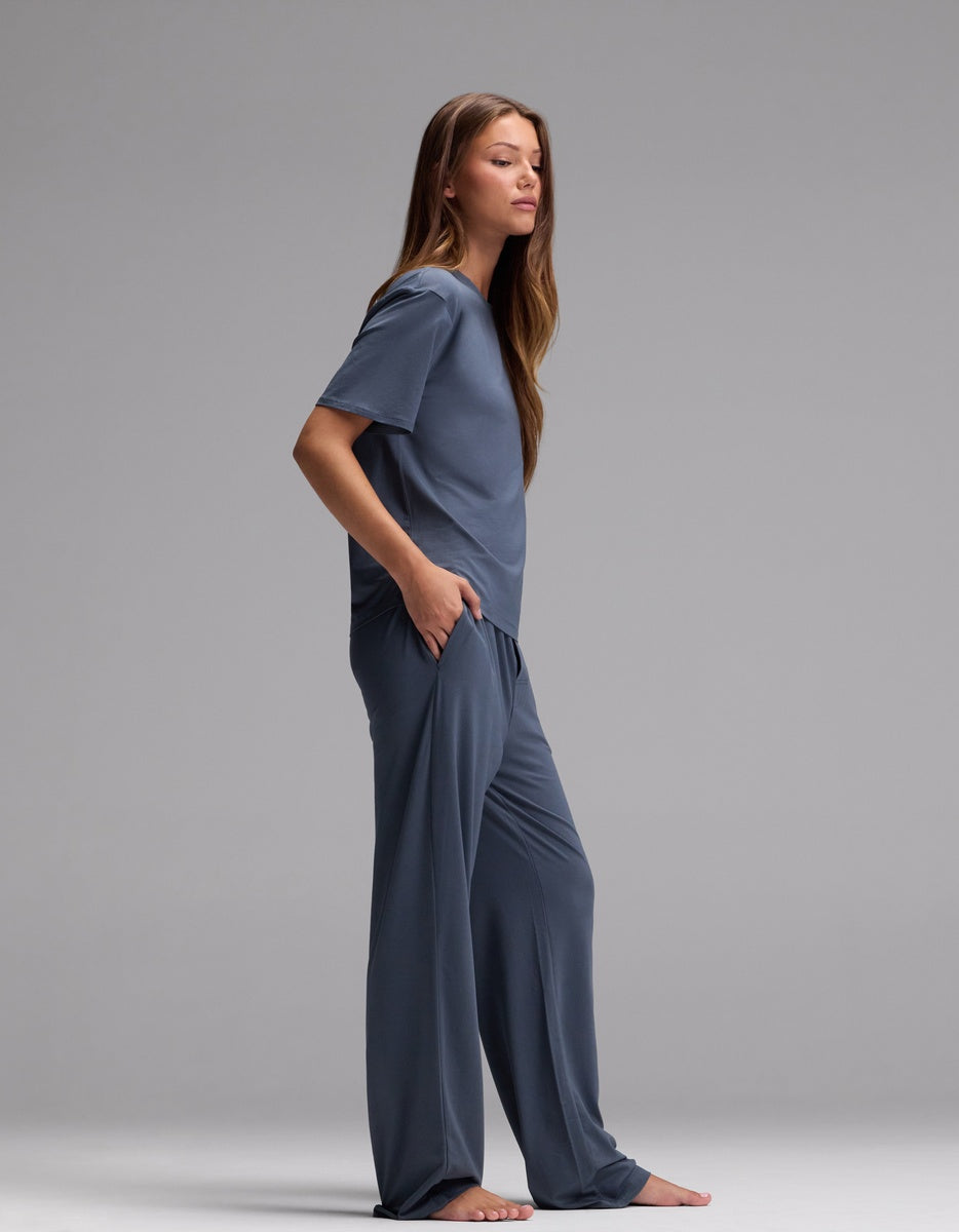 A woman with long brown hair stands barefoot in profile, wearing the Cozy Earth Women’s First Layer Pajama Short Sleeve Tee & Pajama Pant Set in blue, her hands tucked into the pockets of the matching wide-leg pants, against a plain gray background.