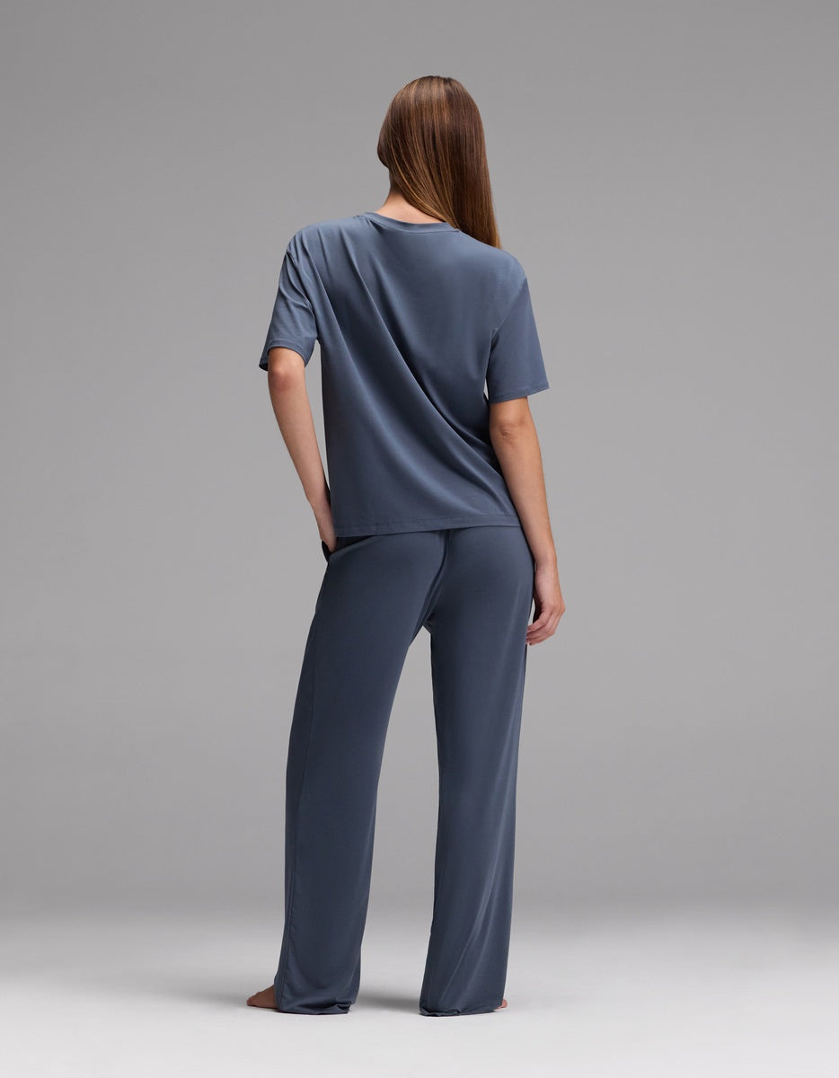 A person with long hair stands barefoot, facing away, wearing the Cozy Earth Women’s First Layer Pajama Short Sleeve Tee & Pajama Pant Set in blue against a plain gray background.