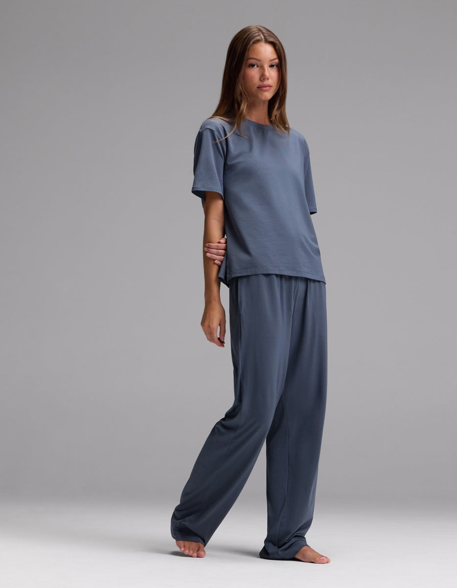 A woman stands barefoot against a plain gray backdrop wearing the Cozy Earth Women’s First Layer Pajama Short Sleeve Tee & Pajama Pant Set in Dawn, featuring a relaxed fit and muted blue hue.