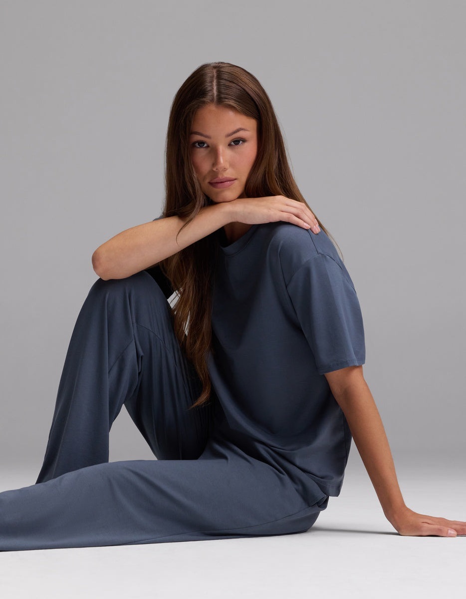 A woman with long brown hair sits on the floor with one arm resting on her knee, wearing the Cozy Earth Women’s First Layer Pajama Short Sleeve Tee & Pajama Pant Set in a loose-fitting blue-gray color, against a plain gray background.