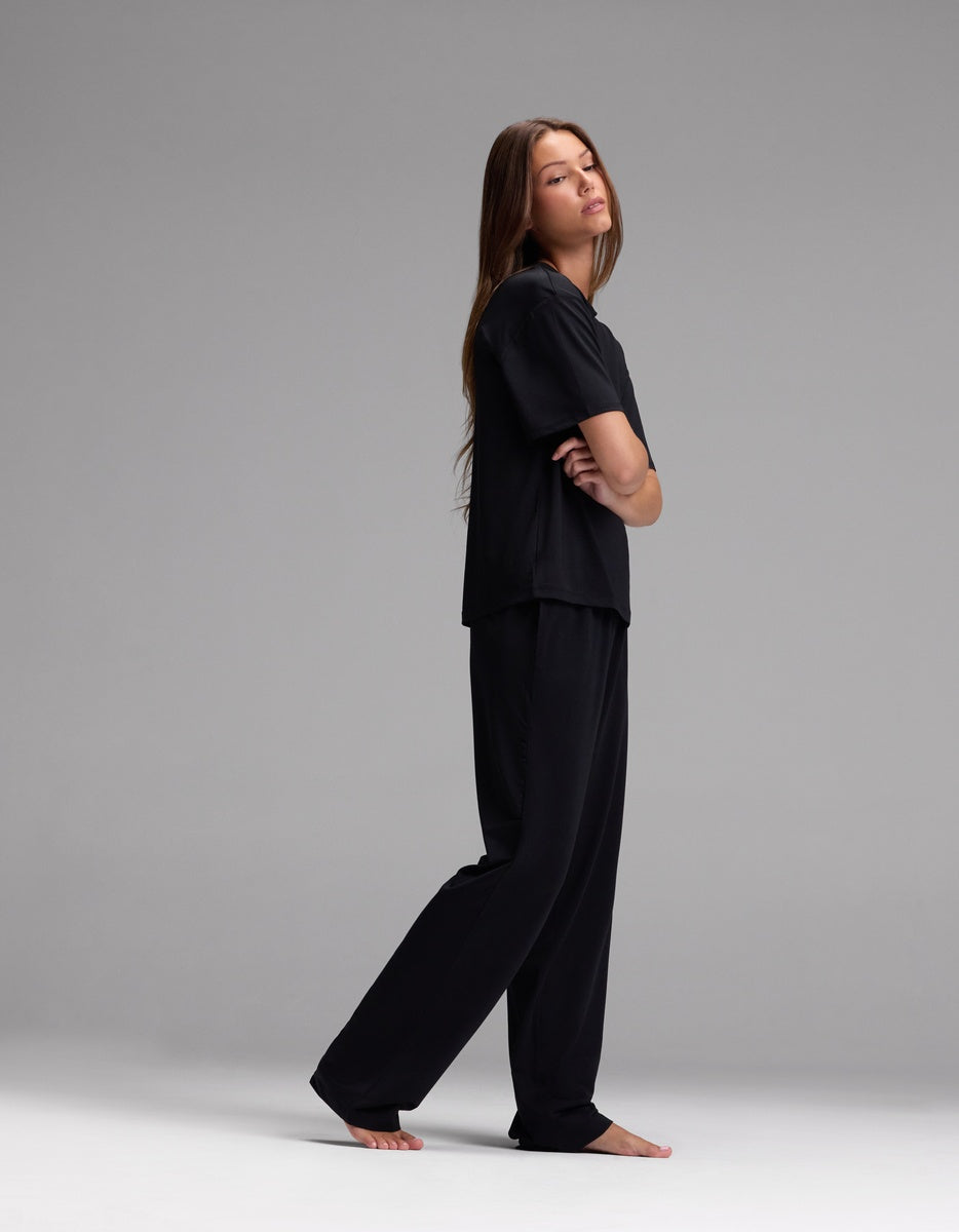 A woman with long hair stands barefoot, posing sideways with her arms crossed against a plain gray background, wearing the Cozy Earth Women’s First Layer Pajama Short Sleeve Tee and Pajama Pant Set in black.