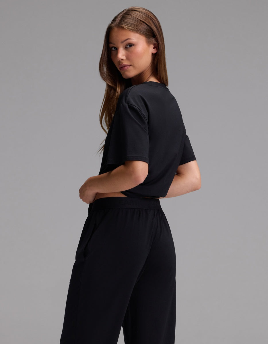 A person with long brown hair stands sideways, looking over their shoulder against a plain gray background while wearing the Cozy Earth Women’s First Layer Pajama Short Sleeve Tee & Pajama Pant Set in black.