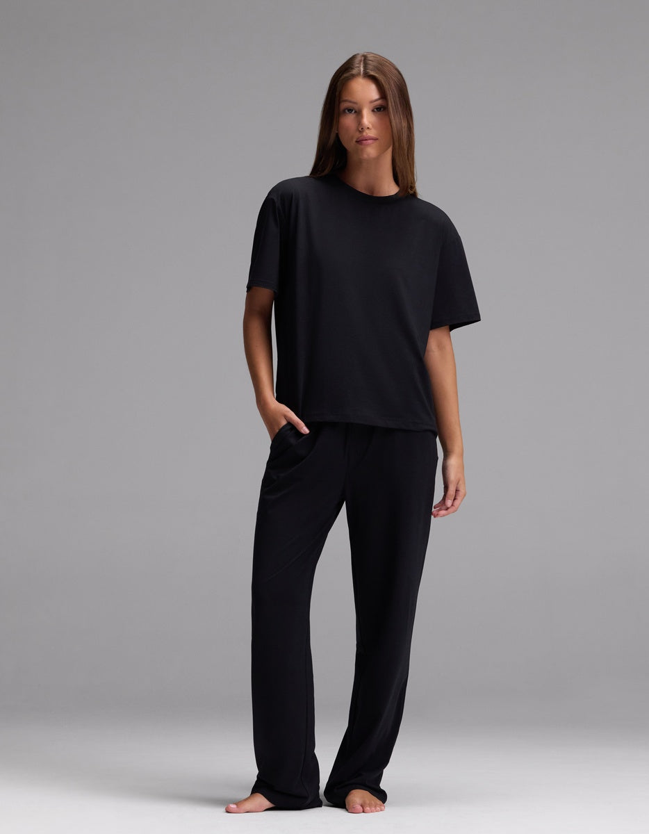 A woman stands barefoot against a plain background, wearing the Cozy Earth Women’s First Layer Pajama Short Sleeve Tee & Pajama Pant Set in Jet Black, with her left hand in her pocket.