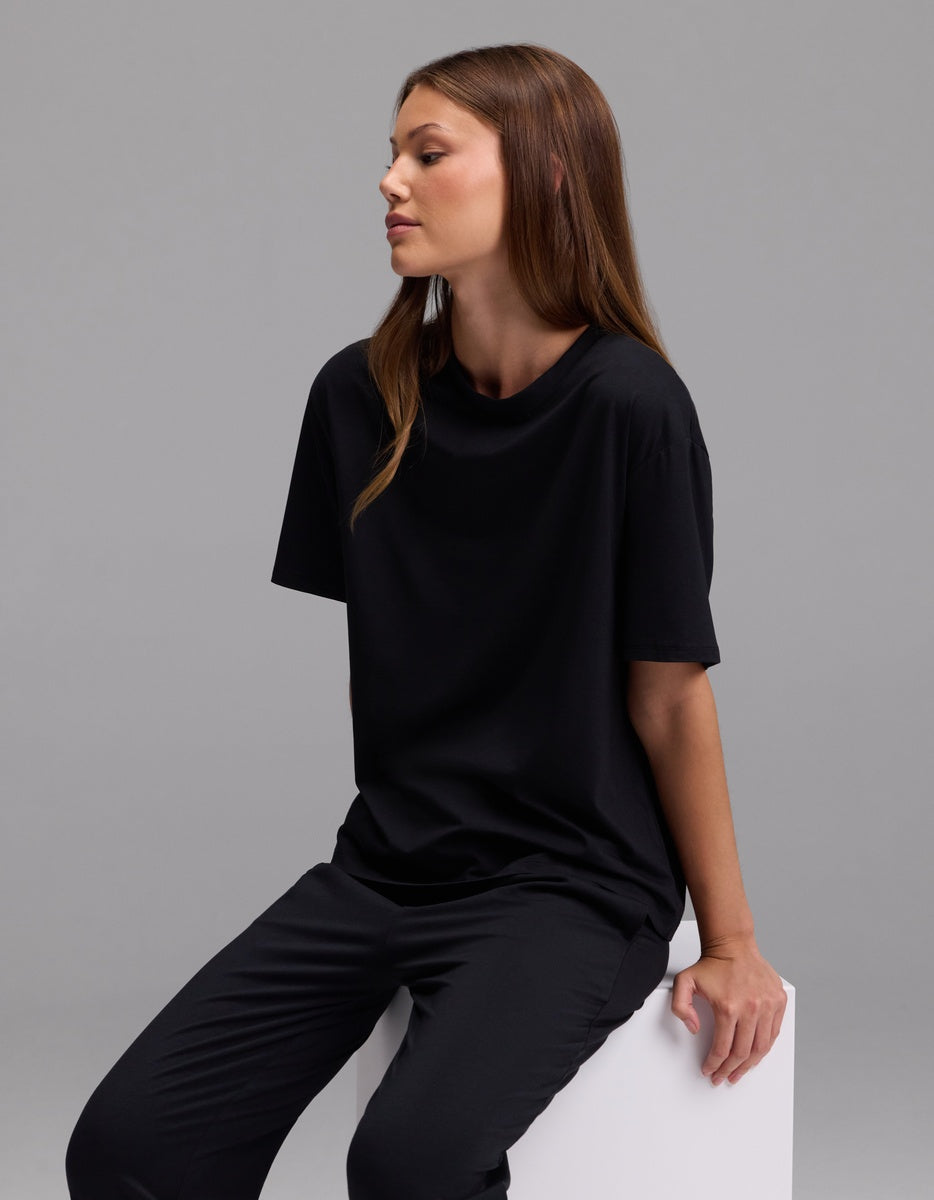 A woman with straight brown hair, wearing the Cozy Earth Women’s First Layer Pajama Short Sleeve Tee & Pajama Pant Set in black, sits on a white block against a plain gray background, looking to the left with a relaxed expression.