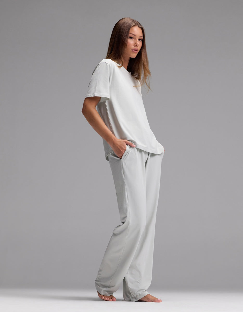 A woman stands barefoot against a gray background, wearing the Cozy Earth Women’s First Layer Pajama Short Sleeve Tee & Pajama Pant Set in light gray, her hands tucked into her pockets as she gazes slightly to the side.