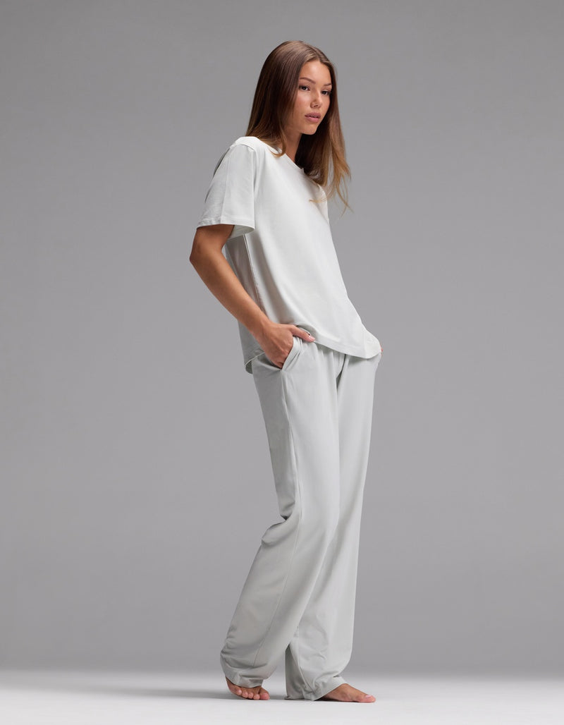 A woman stands barefoot against a gray background, wearing the Cozy Earth Women’s First Layer Pajama Short Sleeve Tee & Pajama Pant Set in light gray, her hands tucked into her pockets as she gazes slightly to the side.