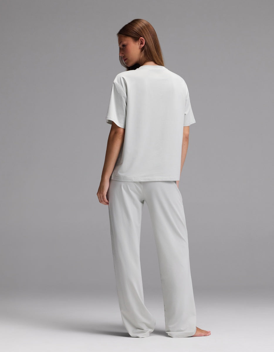 A person stands barefoot on a plain gray background, facing away, wearing the Cozy Earth Women’s First Layer Pajama Short Sleeve Tee & Pajama Pant Set in light gray.