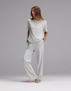 A woman stands barefoot against a gray background, wearing the Cozy Earth Women’s First Layer Pajama Short Sleeve Tee & Pajama Pant Set in Laurel, with her hands in her pockets and looking slightly to the side.|Color:Laurel