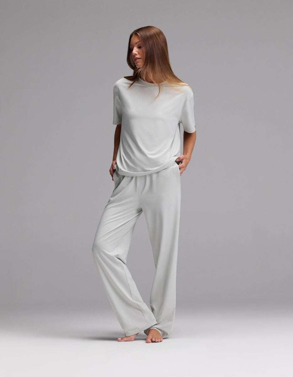 A woman stands barefoot against a gray background, wearing the Cozy Earth Women’s First Layer Pajama Short Sleeve Tee & Pajama Pant Set in Laurel, with her hands in her pockets and looking slightly to the side.|Color:Laurel