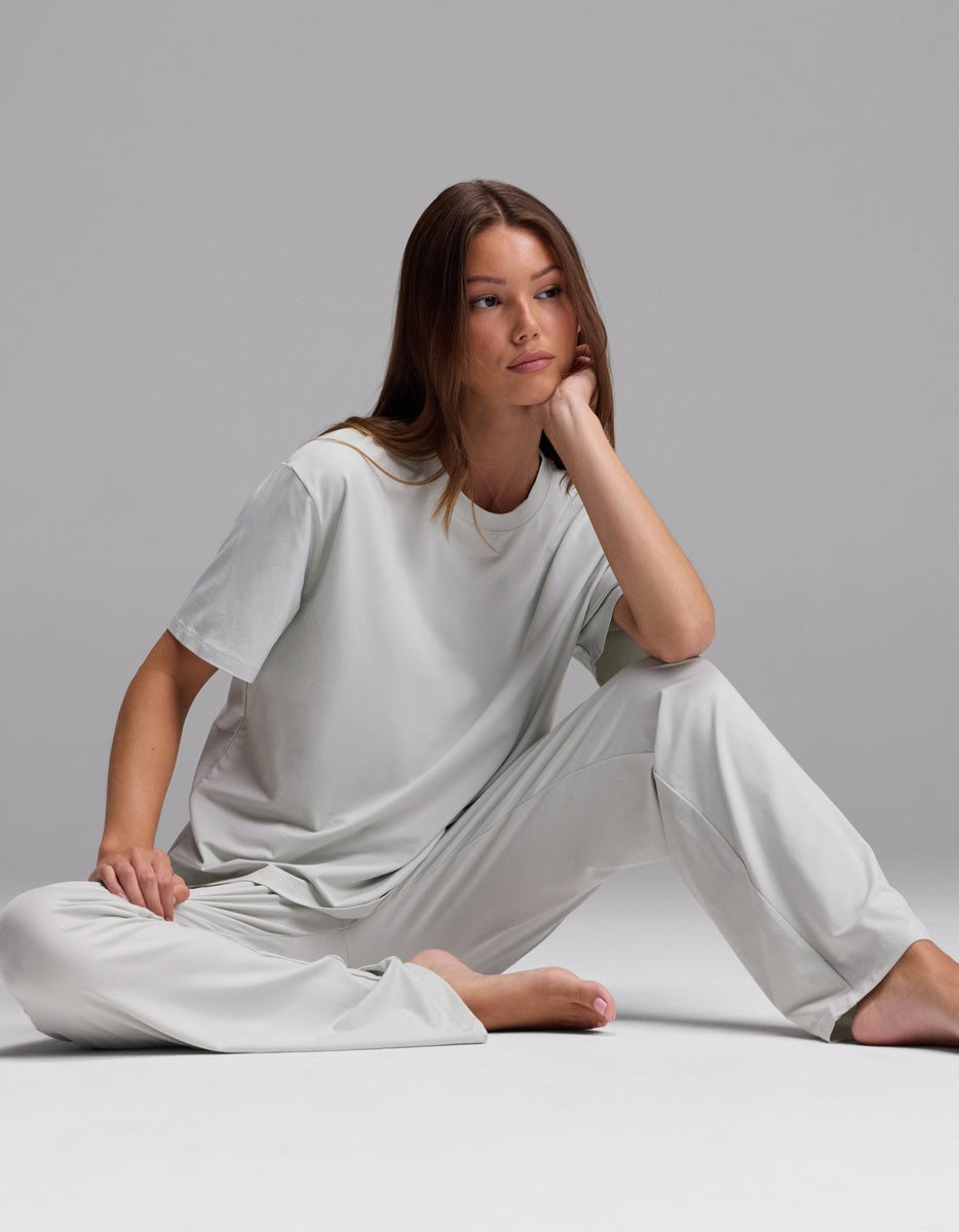 Wearing the Cozy Earth Women’s First Layer Pajama Short Sleeve Tee & Pajama Pant Set in light gray, a woman sits barefoot on the floor against a plain gray background, one arm resting on her knee and a thoughtful expression on her face.