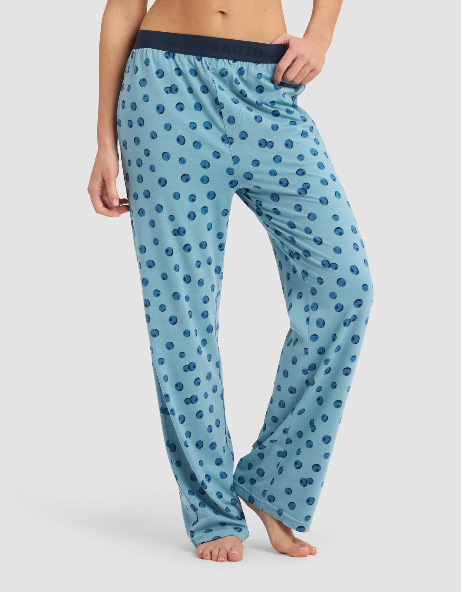 Model is wearing Cozy Earth's Women's First Layer Pajama Pant in light blue with dark blue polka dots and a dark elastic waistband, shown barefoot on a white background.