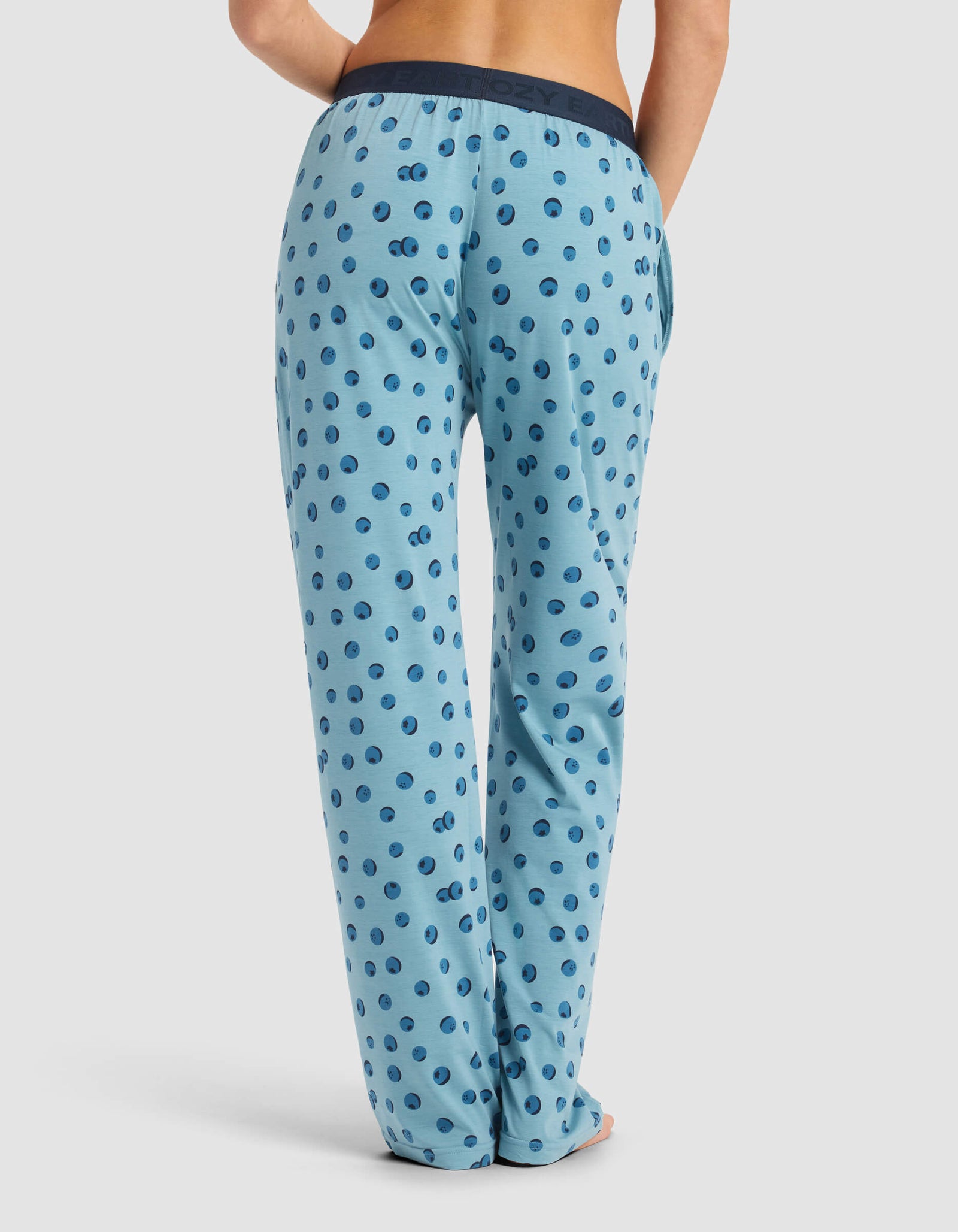 A woman is seen from the back, barefoot on a white background, wearing Cozy Earth’s Women's First Layer Pajama Pant in light blue with dark blue polka dots.