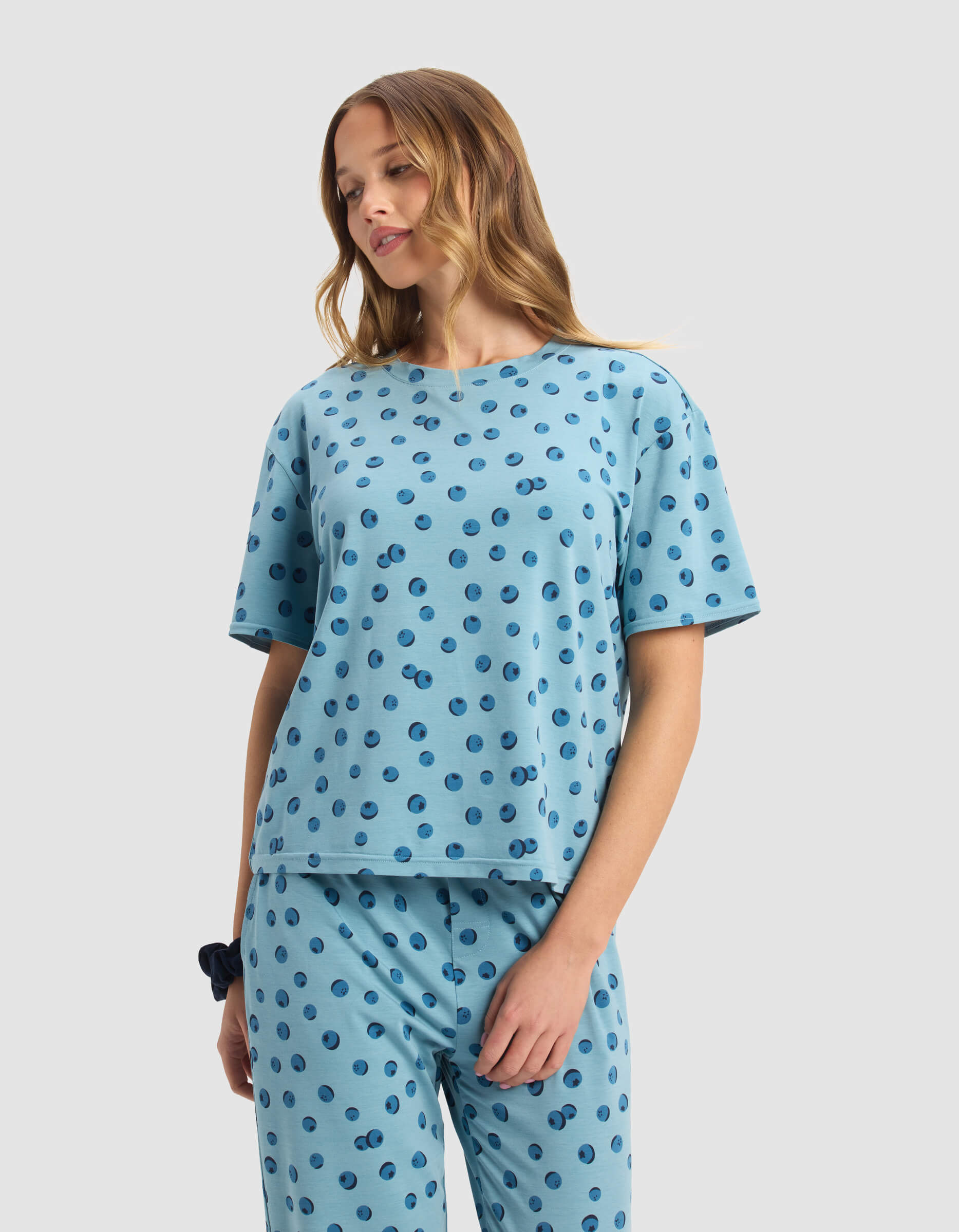 A woman with long, wavy hair stands against a plain light background, looking down in a relaxed mood. She wears Cozy Earth's Women's First Layer Pajama Short Sleeve Tee, a matching light blue set with dark blue polka dots.
