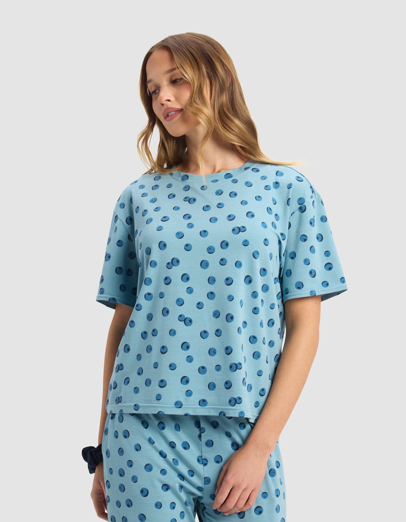 A woman with wavy, light brown hair wears the Cozy Earth Women's First Layer Pajama Short Sleeve Tee & Boxer Short Set in light blue with dark blue polka dots, smiling softly against a plain, light background.