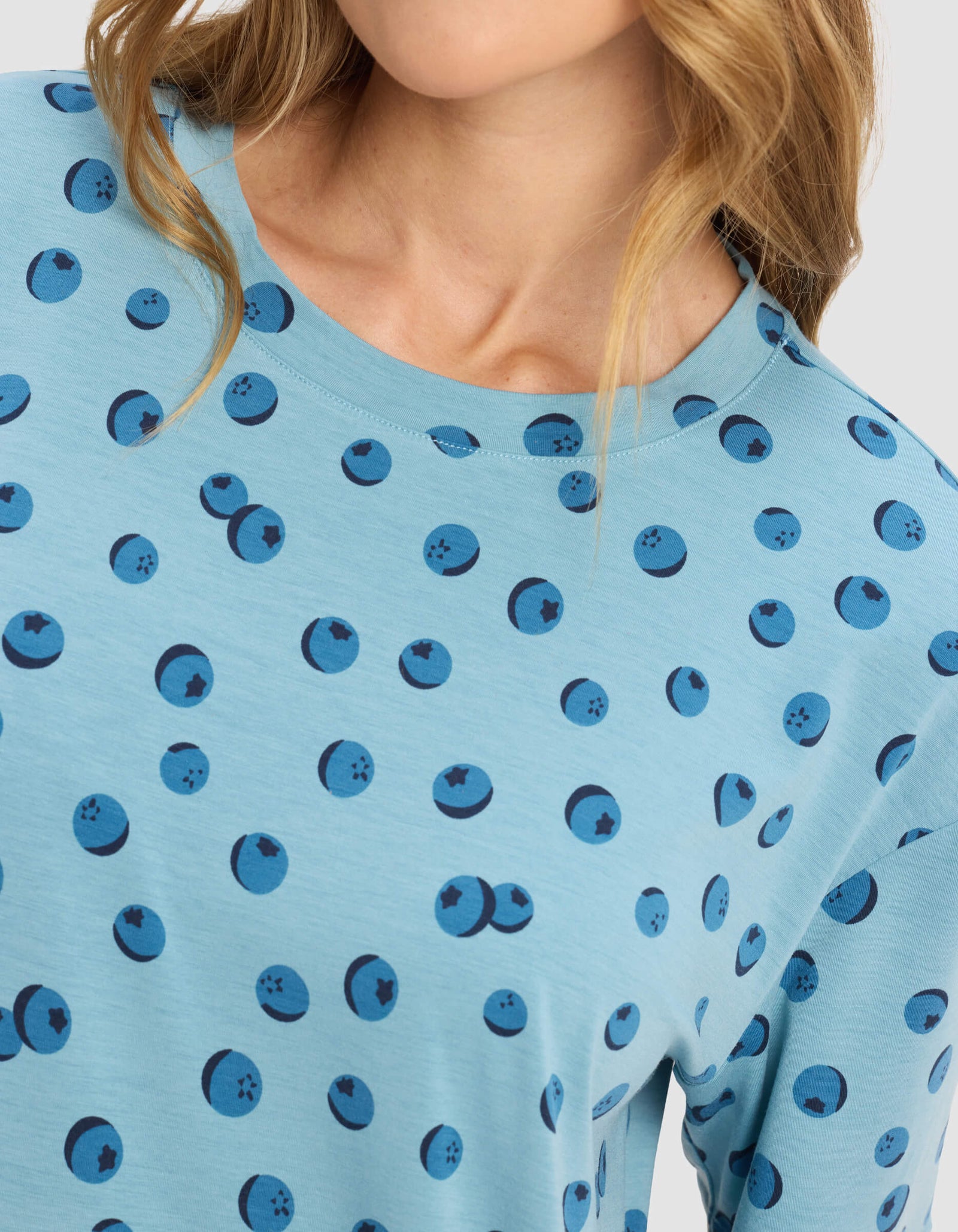 A woman with wavy blonde hair models the Cozy Earth Women's First Layer Pajama Short Sleeve Tee, featuring a light blue base with a blueberry pattern. The image is cropped at her nose and just below her shoulders.