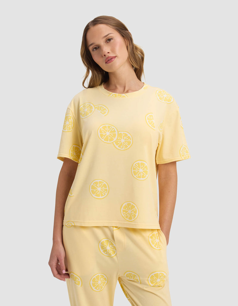 A woman wears the Cozy Earth Women's First Layer Pajama Short Sleeve Tee, paired with matching lemon slice patterned pants, standing against a plain light gray background.
