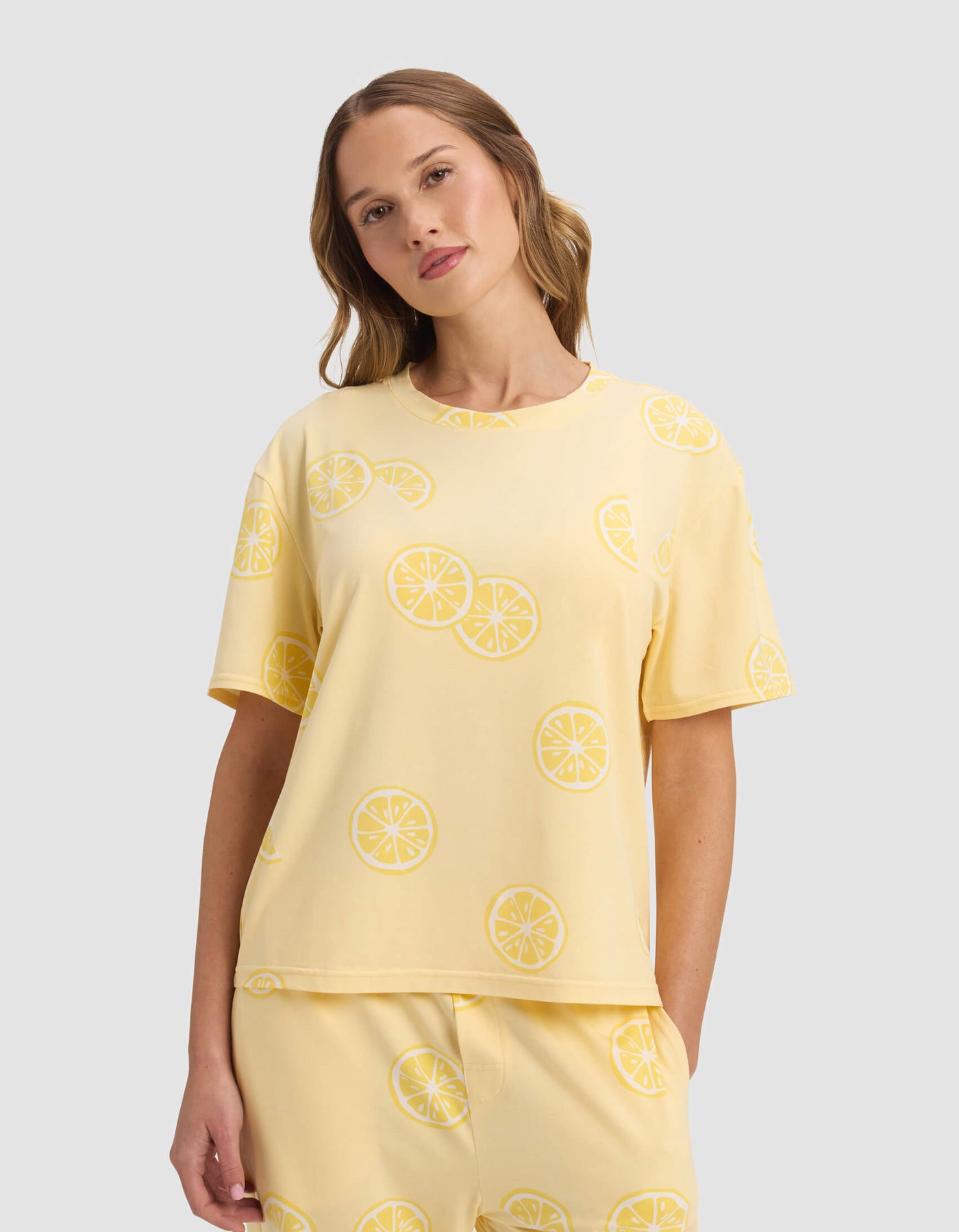 A woman wears the Cozy Earth Women's First Layer Pajama Short Sleeve Tee, paired with matching lemon slice patterned pants, standing against a plain light gray background.